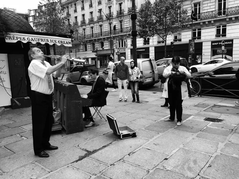 Black-and-white street scene in Paris: a man playing a wind instrument while another plays a street piano.