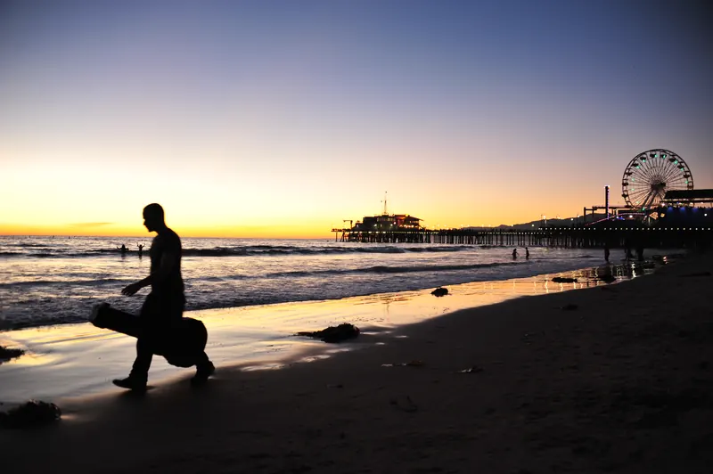 Silhouette of a man carrying a guitar case on Santa Monica Beach at sunset, with the pier behind.